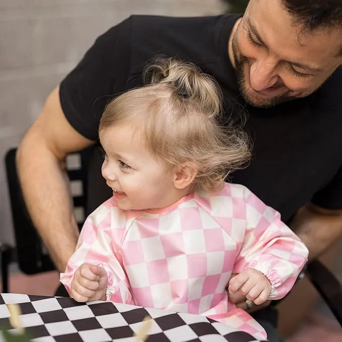 Tiny Twinkle Water Mess Proof Bib in pink checkered pattern worn by a smiling toddler at a table.