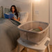 Baby sleeping in mesh bassinet next to bed, mother reading in cozy bedroom setting