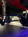Voltz Toys F-450 lifted ride-on car undercarriage close-up showing suspension and wheels indoors.